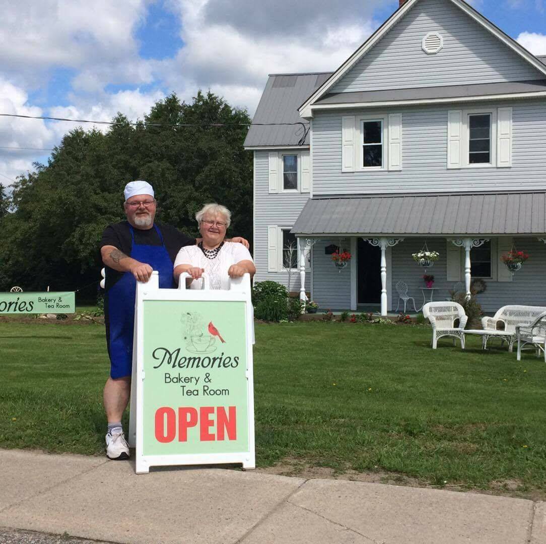 Two people standing by a OPEN sign in front of Memories Bakery and Tea Room