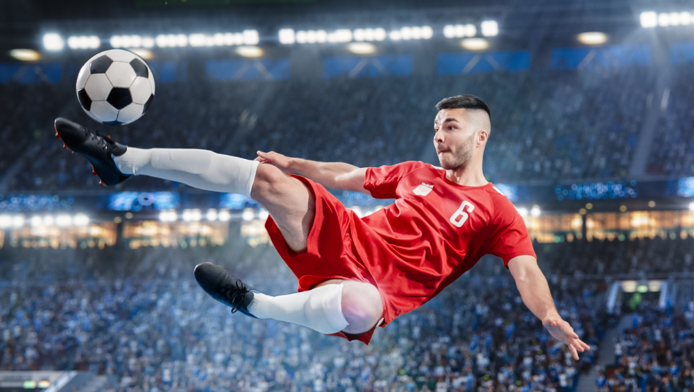 A person kicking a soccer ball in the air. The person is wearing all red and there is a stadium behind them.