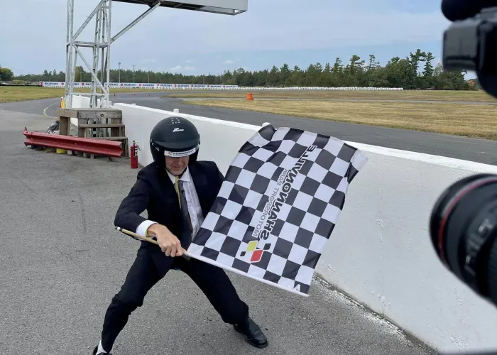 A fellow in a suit and helmet waving a checkered flag next to a race track