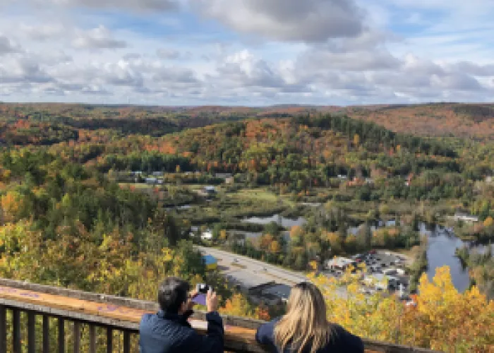 Two people using a lookout, taking a picture of a town below with beautiful fall colours in the distance.