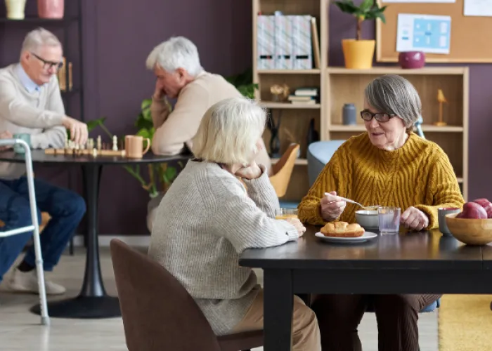 Retirement residence. Two women sitting at a table eating and two men in the background playing chess. The walls are purple with bookshelves.
