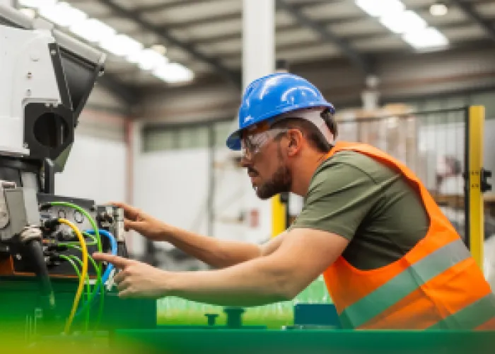 A trades person working on a machine. The person is wearing a blue hard hat and orange safety vest in a warehouse or factory.
