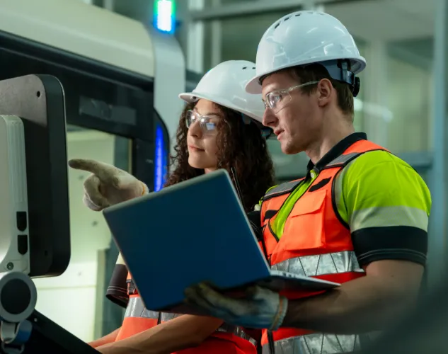 People working on a machine. One person is holding a laptop the other is at the controls. Both are wearing hard hats, safety glasses, safety vests. 